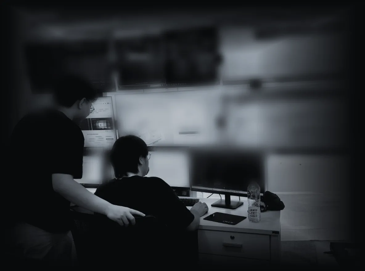 Two Cyber Defense analysts working at a desk in the Bangkok SOC, a wall of consoles behind them, the screens deliberately out of focus.